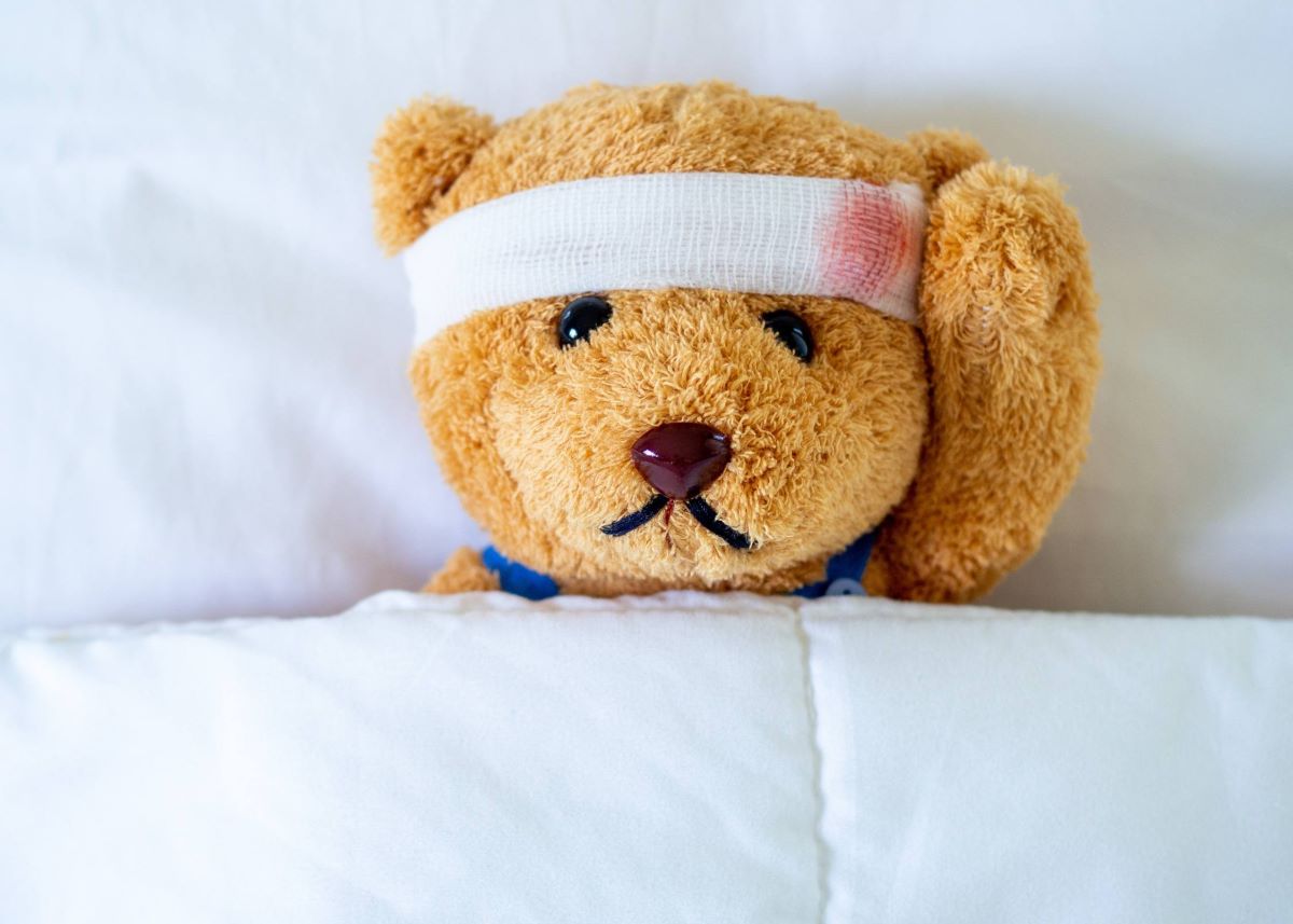 Teddy bear lying in bed with gauze wrapped around its forehead